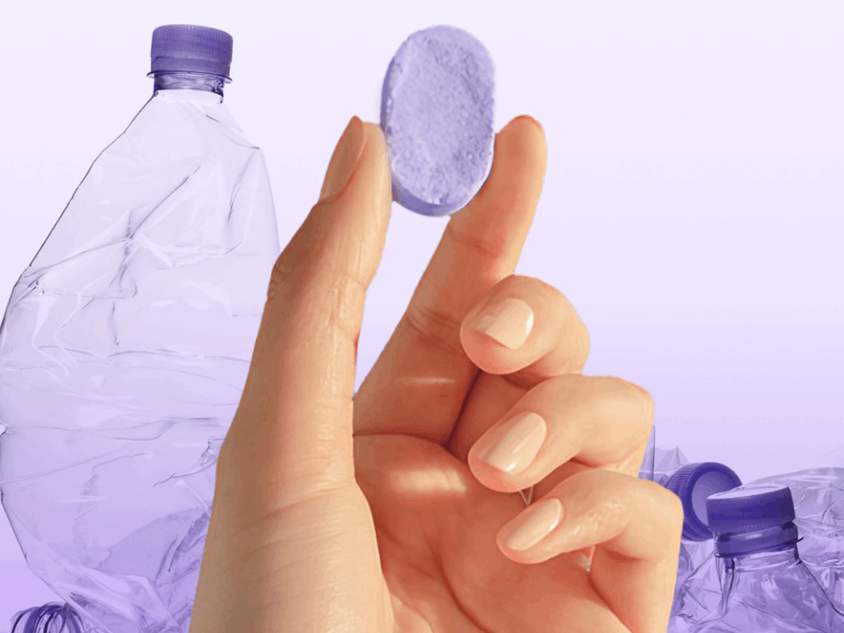 Concept visual showing a hand holding one purple cleaning tablet in front of translucent single-use plastic bottles on a lavender studio background