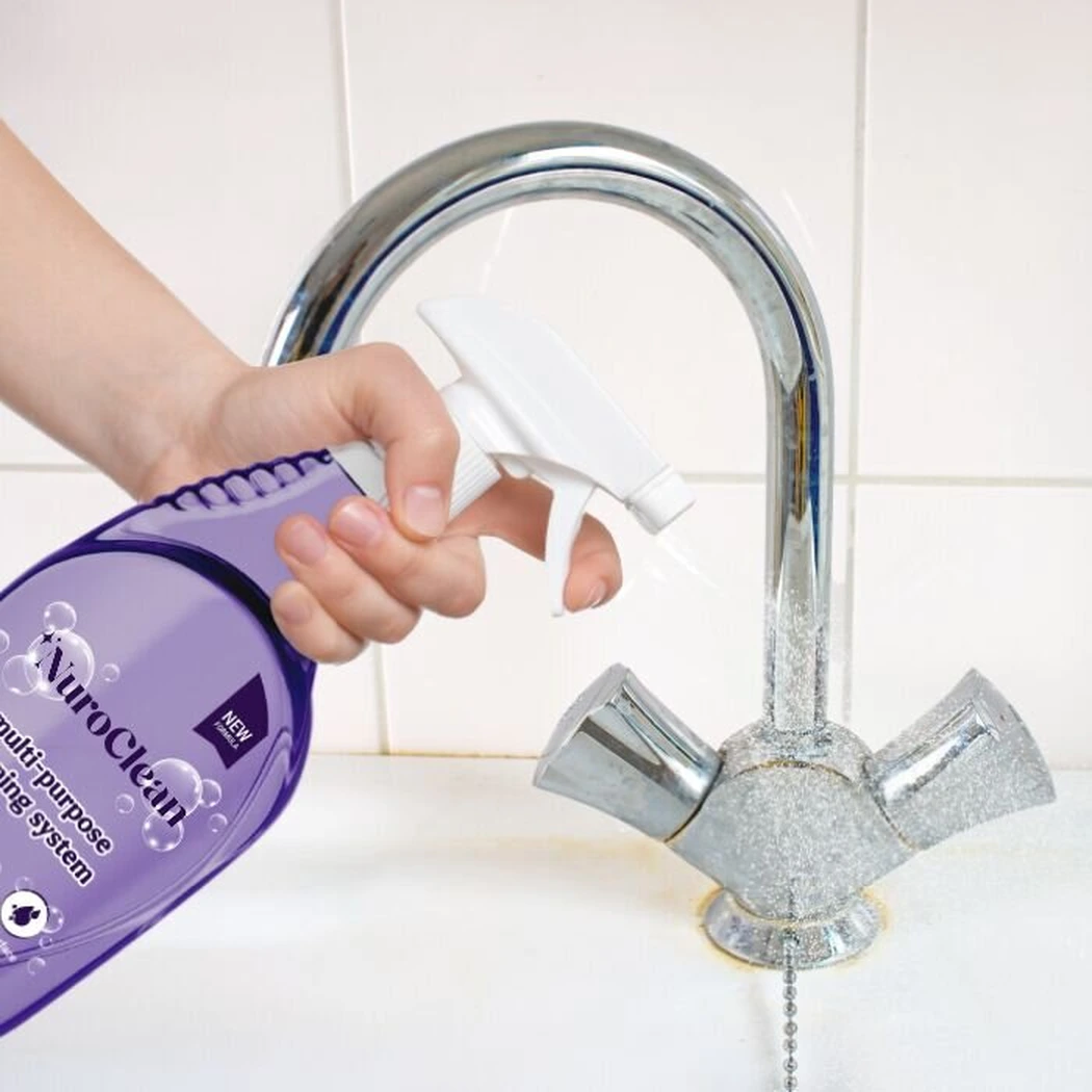 Hand holding the NuroClean spray bottle aimed toward a clean chrome kitchen faucet and white ceramic tiled counter